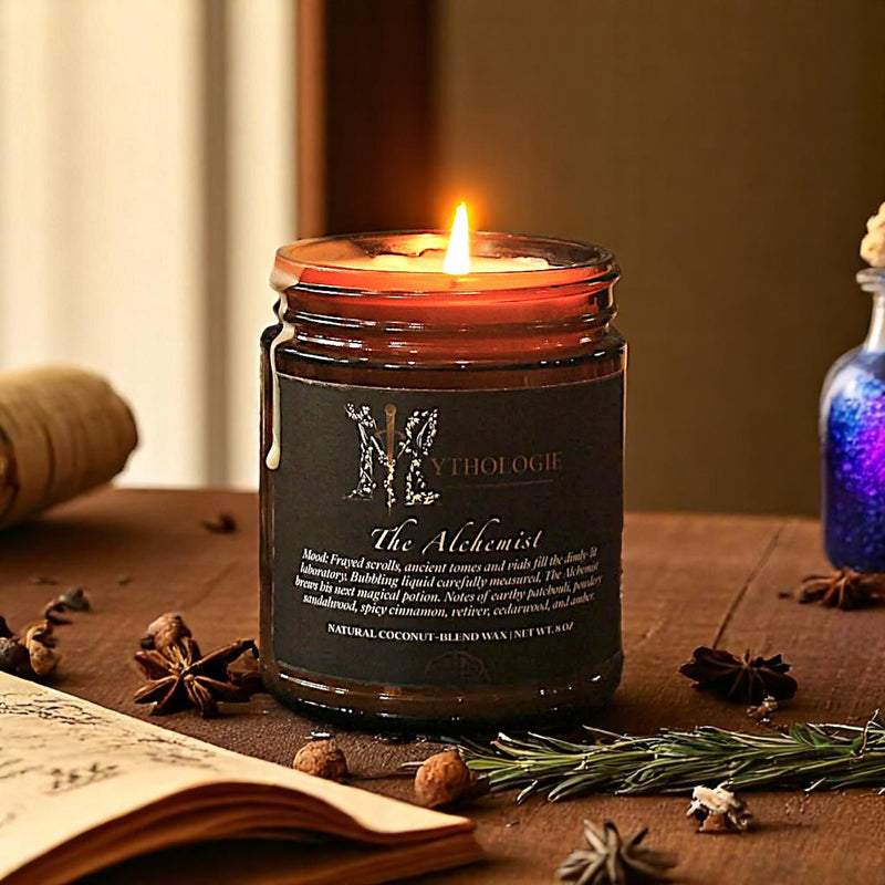 A red candle with a black label, titled 'The Alchemist', burning on a table with lab equipment and old books in the background, creating a cozy, historical ambiance.